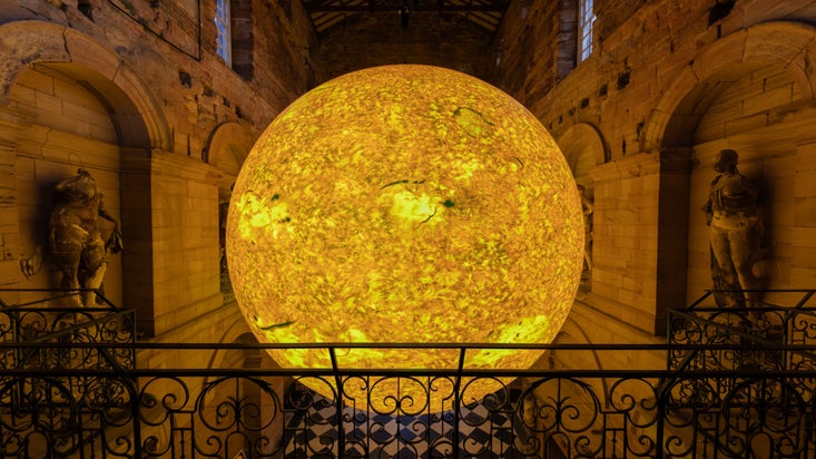 A large, scaled sculpture of the sun sits suspended inside the centre of an empty room. Beneath it, there is a black and white checked floor. The shot is taken from a balcony, with a black railing in front of the sun.